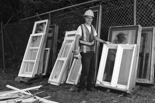 A male worker wearing a safety helmet, high-visibility vest, and dark trousers stands outdoors on a grassy area near a chain-link fence. He is holding a white-framed window with glass panes, which appears to be part of a collection of similar windows leaning against the fence and stacked on the ground. The windows vary slightly in size but share a consistent white finish and clear glass. The background includes additional windows and framing materials set against the fence, with trees or foliage visible behind the fencing. The scene is evenly lit, suggesting natural daylight, and the setting appears to be a designated area for rubbish removal or disposal related to office or property clearance, consistent with services offered by Office Clearance Finsbury Park.