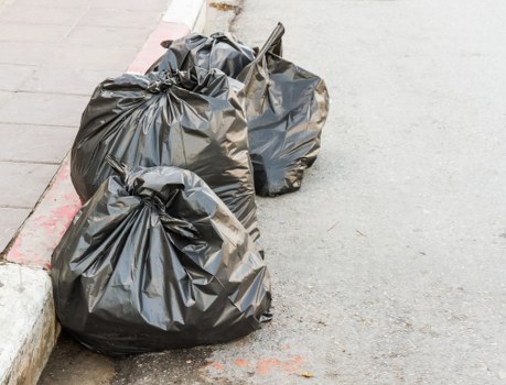 Two large black plastic rubbish bags filled with waste are placed on a grey paved sidewalk next to a curb with a red and white painted edge, likely part of a residential or commercial area in Finsbury Park. The bags are slightly crumpled and bulging, indicating they've been filled with general household or office waste. One bag is leaning against a metal tool, possibly a rake or broom, which is partially visible and resting on top of a bag. The background shows an uneven concrete street surface with some faint markings and a small section of the street perimeter, suggesting this scene is located in an urban setting where rubbish collection services like those from Office Clearance Finsbury Park might be operating. The environment appears clean, with no visible debris or additional waste nearby, emphasizing the typical appearance of waste ready for collection within the local area.