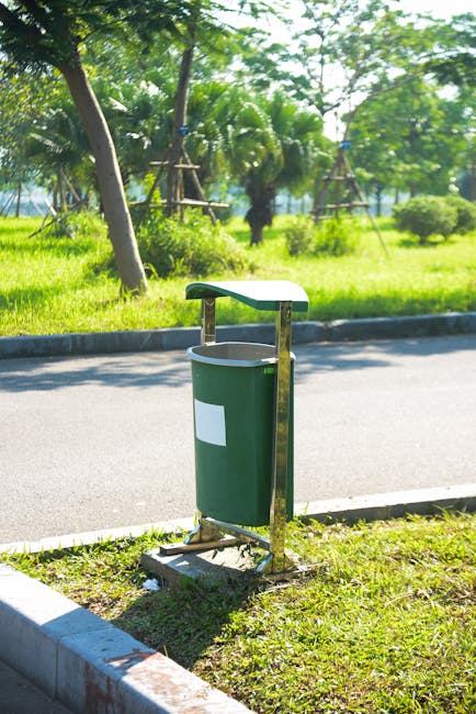 A cylindrical metal trash bin with a rounded top and a wide open slot at the top for disposing of waste, standing on a metal post amidst dense green foliage of trees and bushes. The bin displays a bla