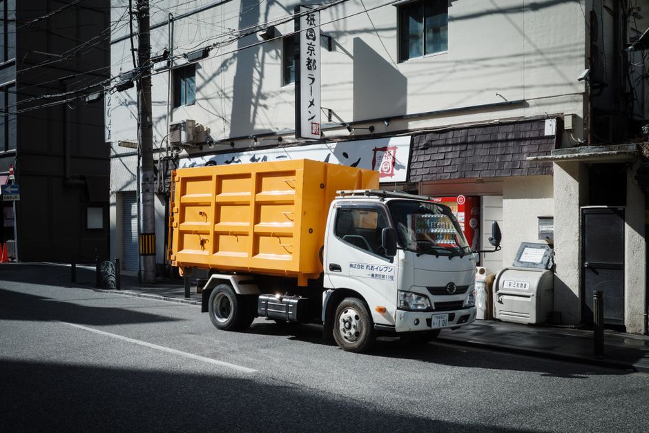 A large commercial rubbish collection truck is parked on a city street with multi-storey residential and office buildings in the background. The truck features a white cab with a silver fuel tank and 