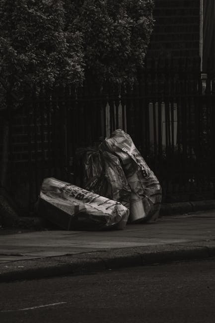Three large black plastic trash bags filled with waste are placed on the pavement beside a black metal fence, with one of the bags partially leaning against a small, leafy green bush. The bags appear